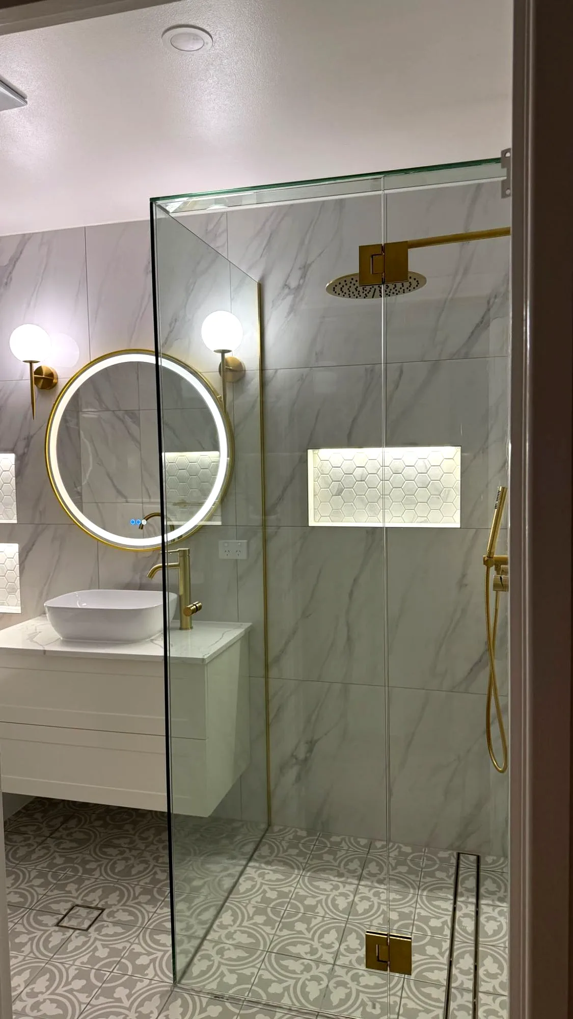 Marble-look bathroom with a frameless glass shower, brass fittings and a round LED mirror