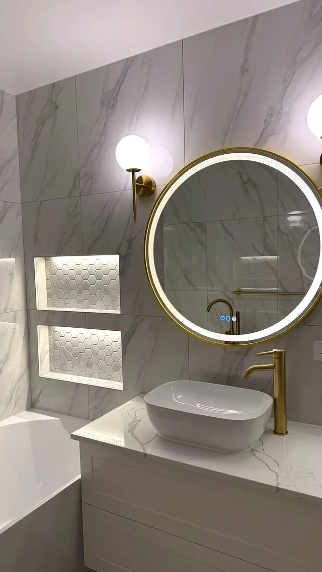Round LED mirror with gold frame above a white basin and brass tap on a marble-look benchtop